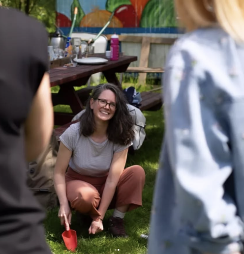 Vrouw lacht terwijl ze met een rode schep hurkt bij een picknicktafel in een zonnige tuin.