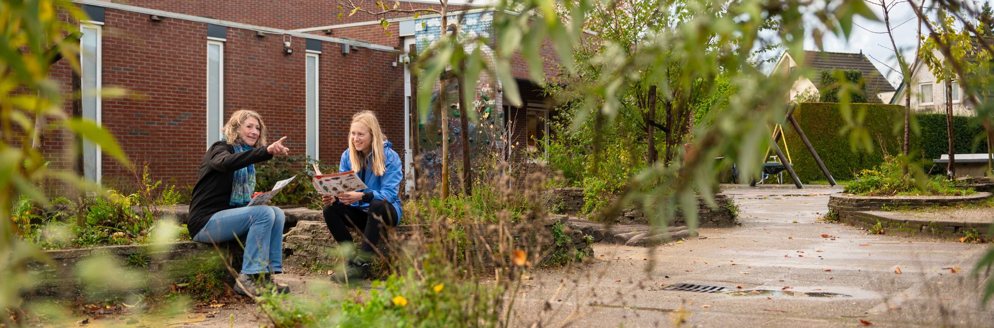 Twee mensen zittend in tuin bij gebouw, lezend en pratend met planten en schommel op achtergrond.