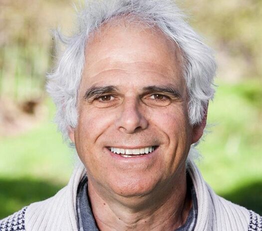 Een oudere man met wit haar glimlacht in een groene, wazige buitenomgeving.