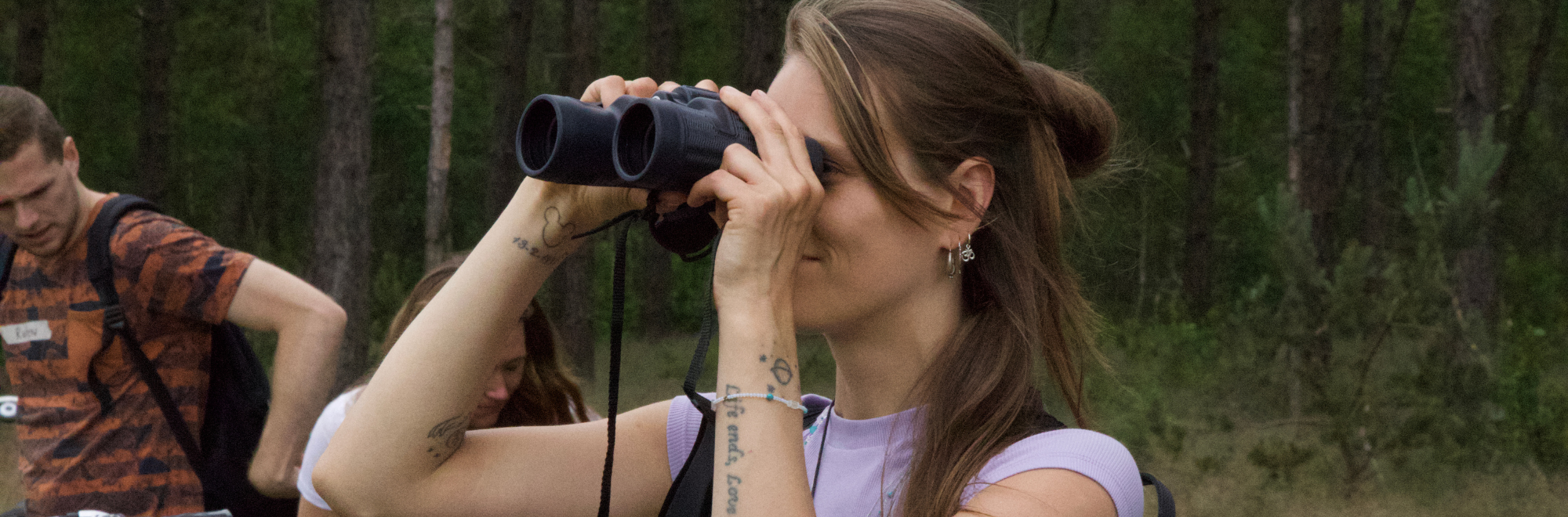Een vrouw kijkt door een verrekijker in een bos, omgeven door bomen.