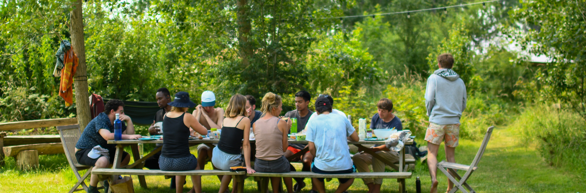 Groep mensen eet samen aan een picknicktafel in een groene, bosrijke omgeving.