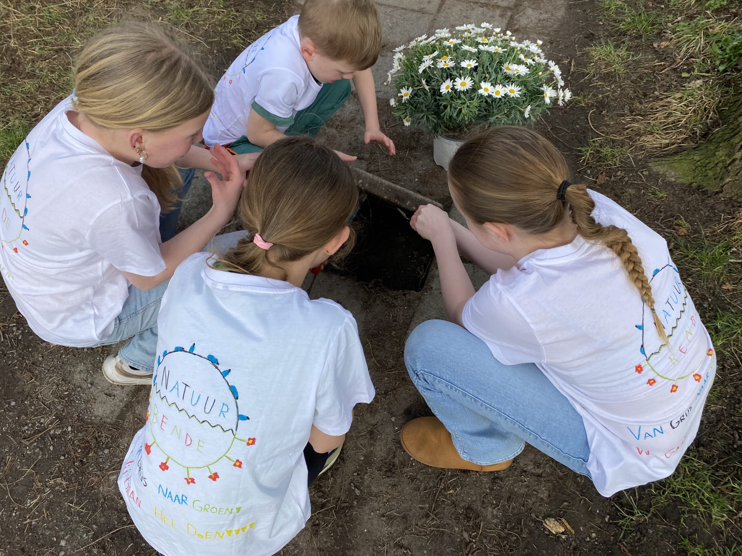 Kinderen in witte T-shirts onderzoeken een gat in de grond naast een pot witte bloemen.