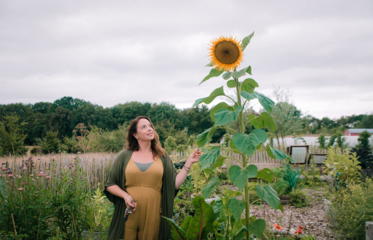 Vrouw in tuin wijst naar hoge zonnebloem, met groene achtergrond en bewolkte hemel.
