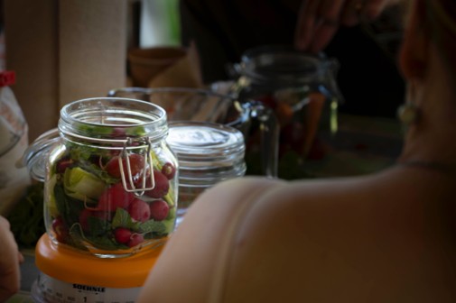 Pot met felgekleurde radijsjes en slablaadjes, omringd door glazen potten en een persoon op de voorgrond.