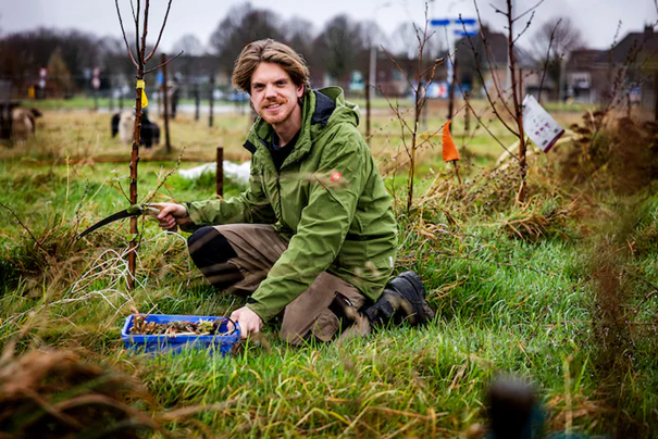 Persoon knielt in grasland, snoeit jonge boom en lacht naar de camera, met schapen op de achtergrond.