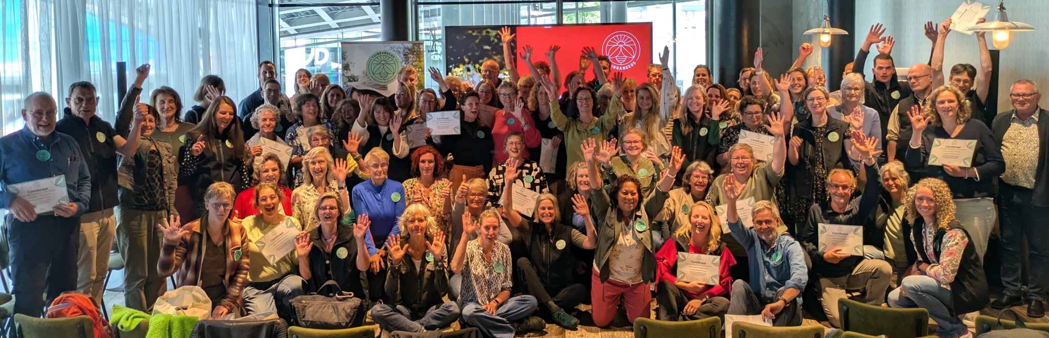 Grote groep mensen viert met certificaten en opgeheven handen in een lichte zaal.