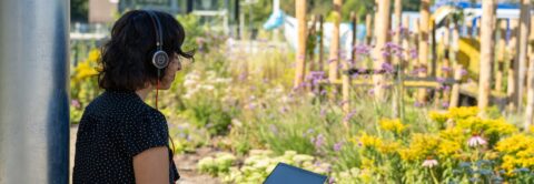 Vrouw met koptelefoon leest op tablet in een kleurrijke tuin vol bloemen.