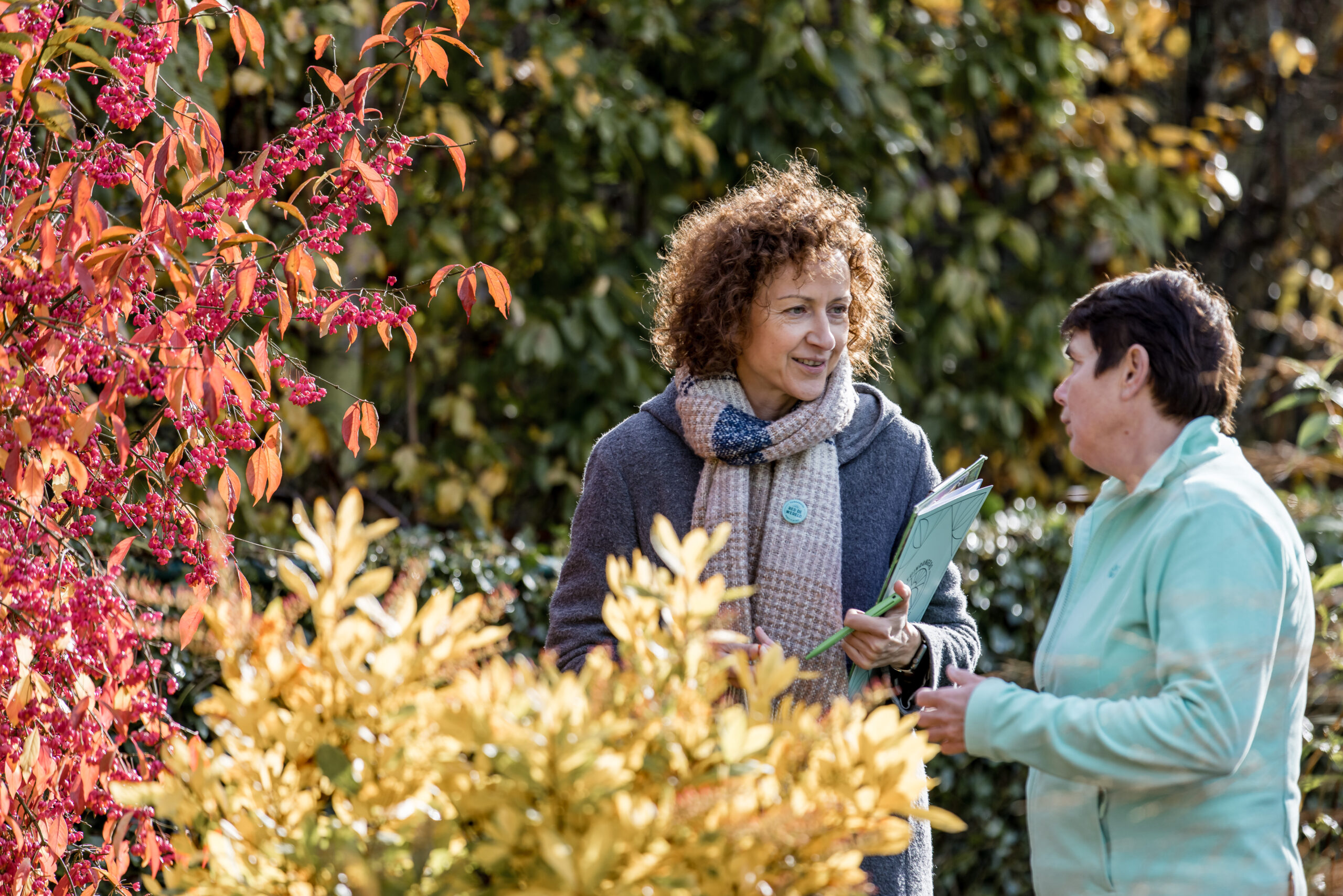 Twee vrouwen staan buiten in een tuin met kleurrijke herfstbladeren, pratend en lachend. De ene houdt folders vast en draagt een sjaal en jas; de andere draagt een lichte jas.