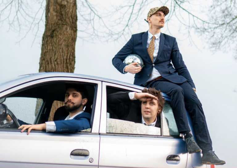 Drie mannen in pakken op en in een auto; een man houdt een discobal vast, een ander tuurt in de verte.