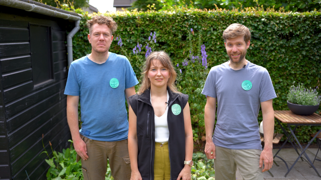 Drie mensen staan buiten in een tuin en glimlachen naar de camera. Ze dragen elk een ronde groene badge. Op de achtergrond staan groene heggen, bloemen en een houten tafel met potplanten.