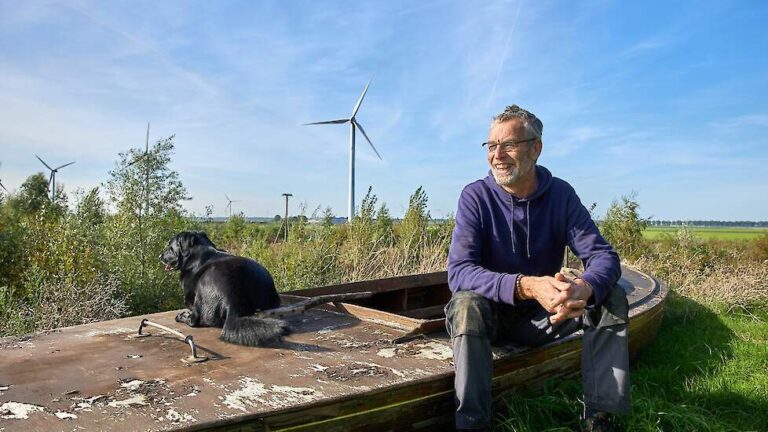Man en hond op een oude boot voor een veld met windmolens en blauwe lucht.