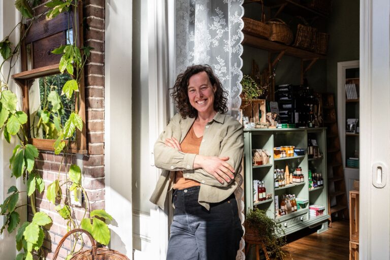 Vrouw glimlacht in een winkel met groene bladeren en producten op een plank.