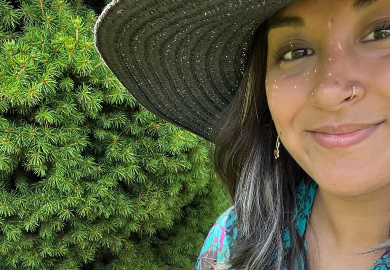 Vrouw met hoed glimlacht naast groene struik.