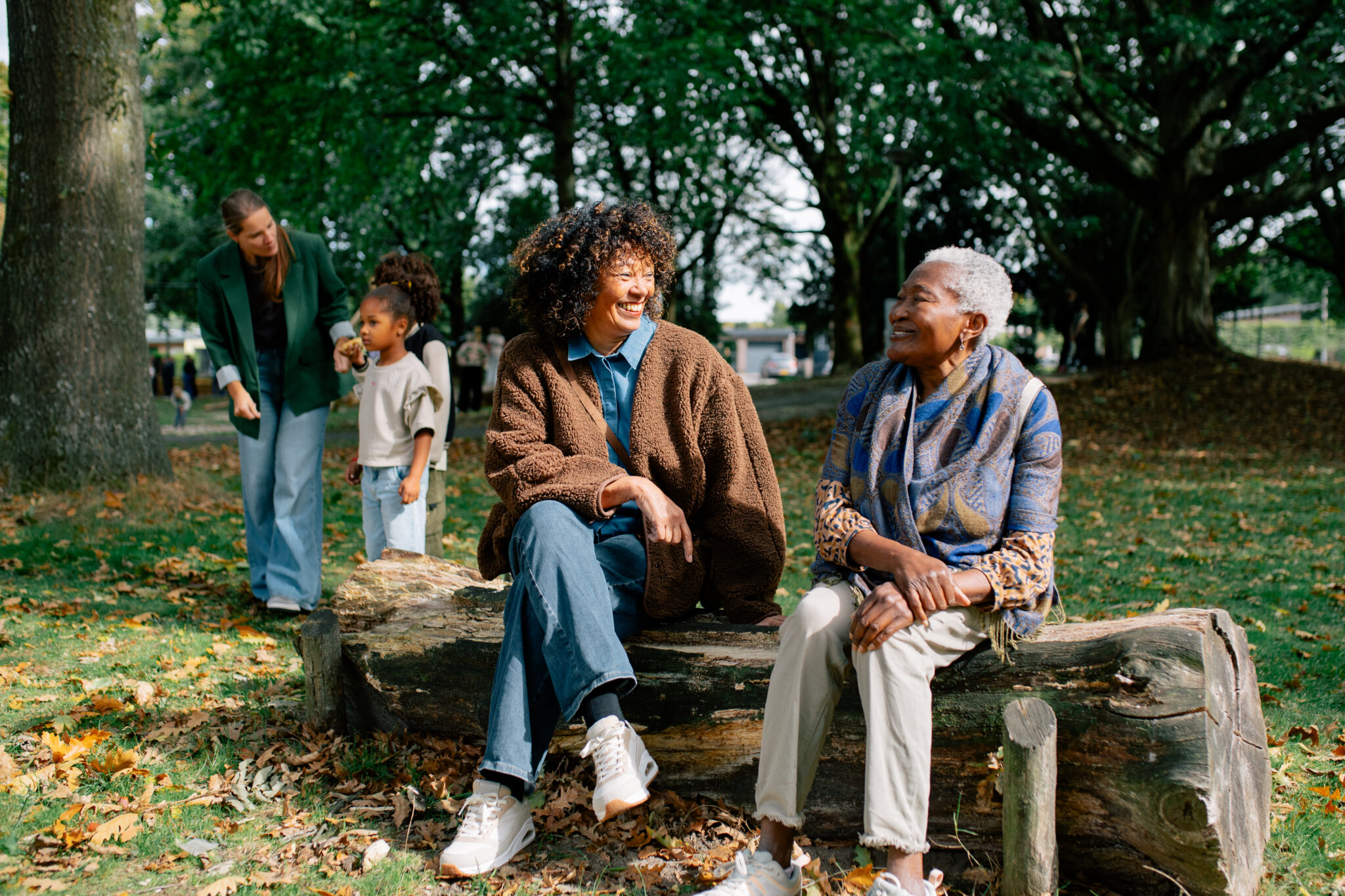 Twee vrouwen zitten op een boomstam in een park, lachend en pratend, met kinderen op de achtergrond.