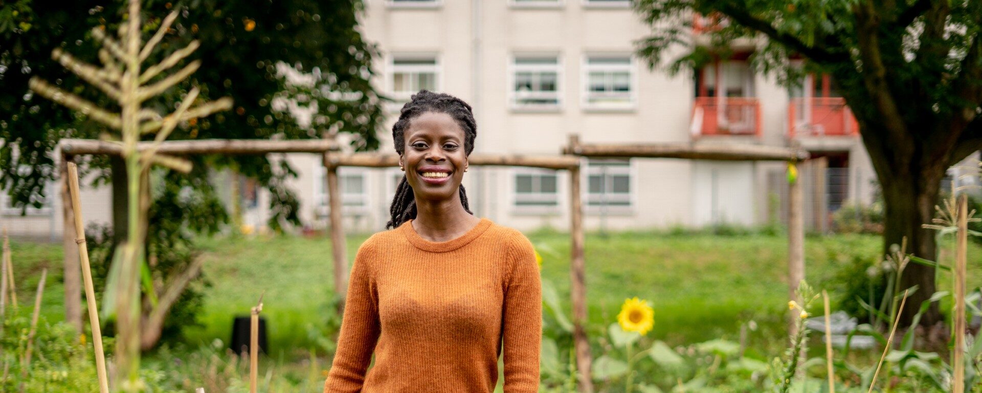 Vrouw in oranje trui glimlacht in een tuin met zonnebloem, gebouw op achtergrond.