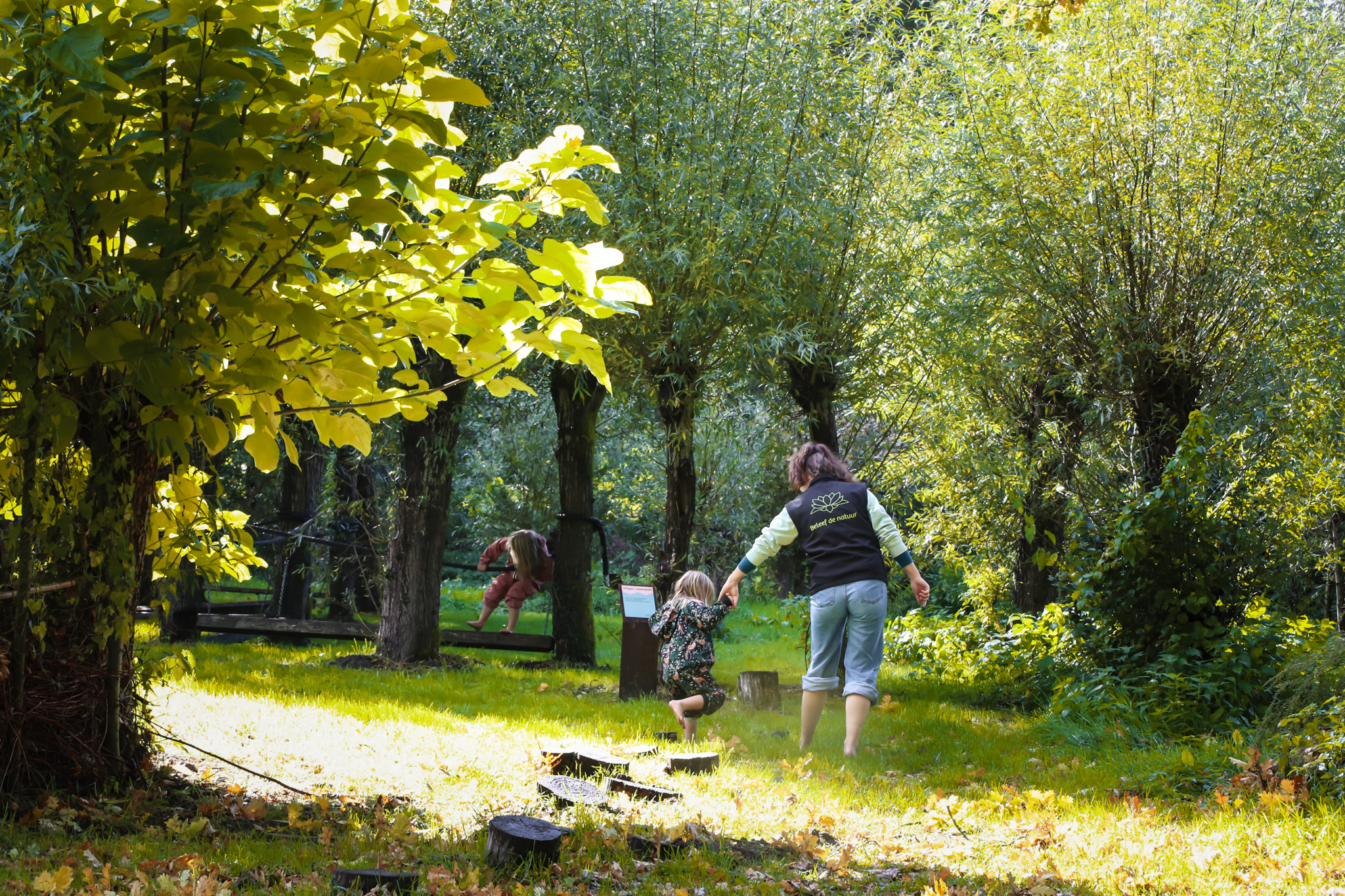 Twee mensen lopen hand in hand door een bosrijke tuin met zonlicht en groene bomen.