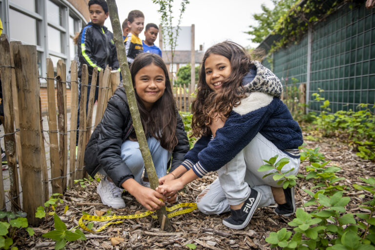 Twee meisjes planten samen een boom in een tuin, omringd door andere kinderen.