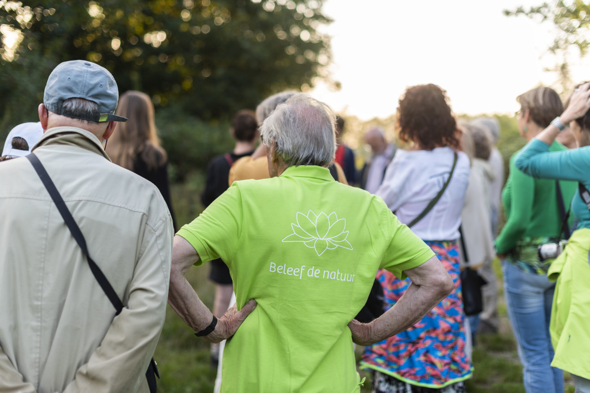 Groep mensen in een bos, persoon in groen shirt met "Beleef de natuur" erop gedrukt.
