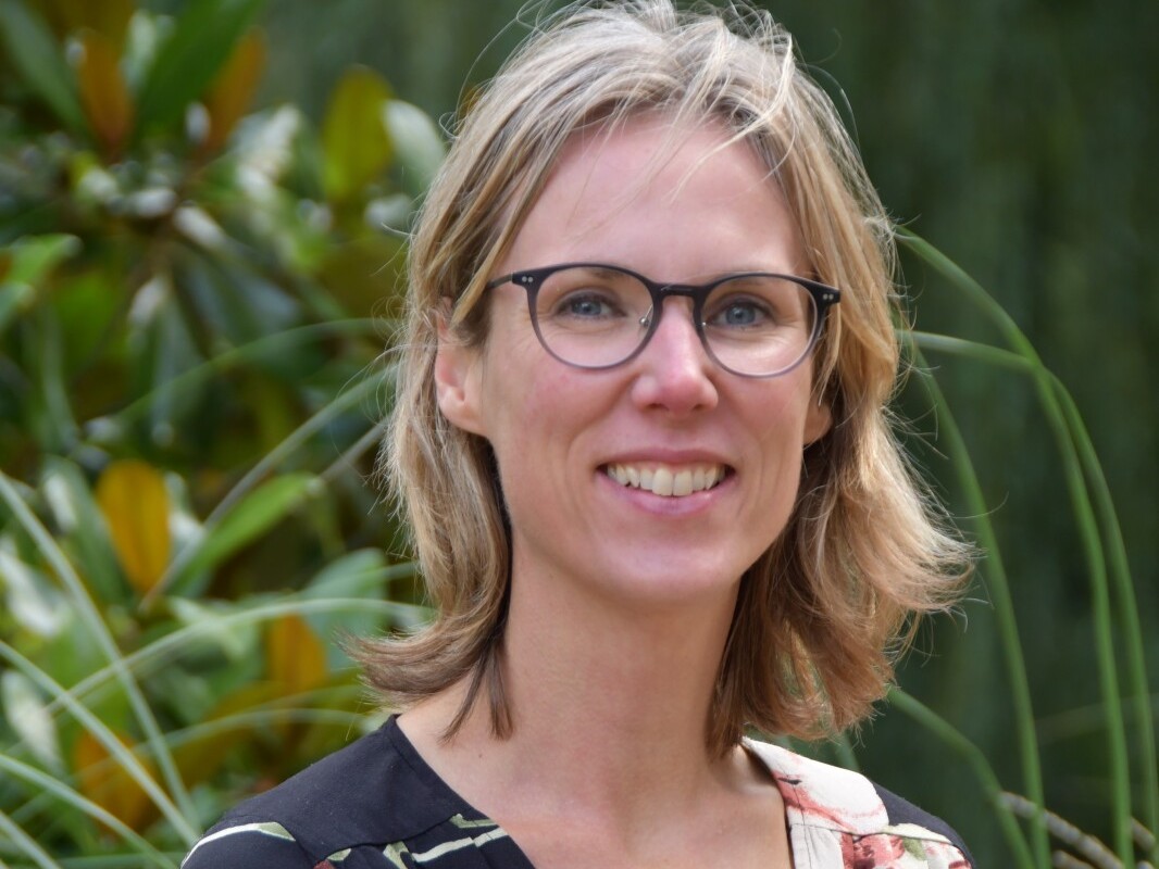 Een vrouw met bril glimlacht buiten, omringd door groene planten.