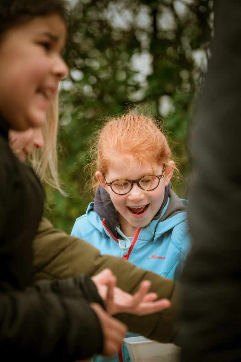 Een lachend kind met bril en blauwe jas kijkt naar handen tijdens een buitenactiviteit.