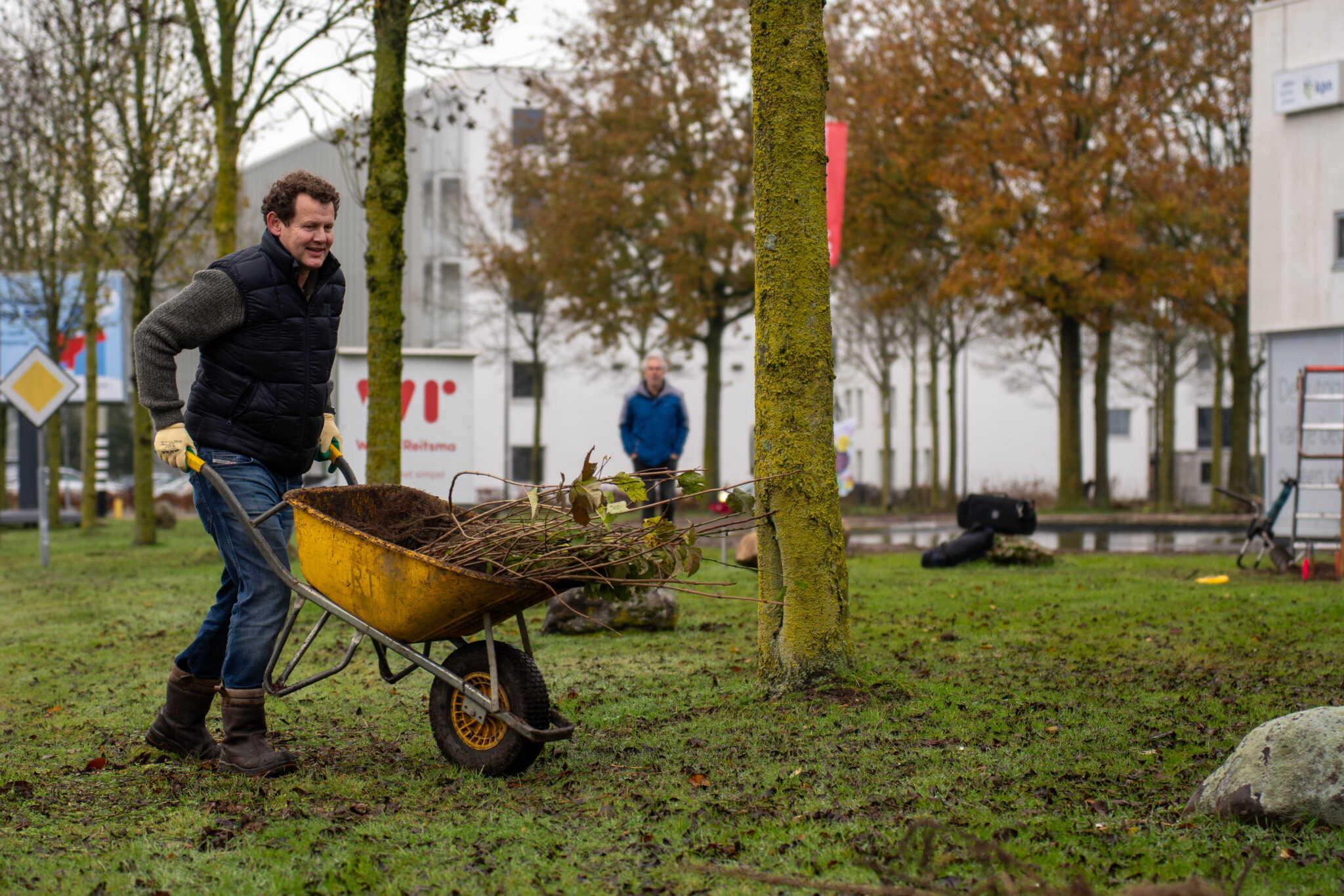 Man duwt kruiwagen vol takken over grasveld, omringd door bomen en gebouwen op achtergrond.