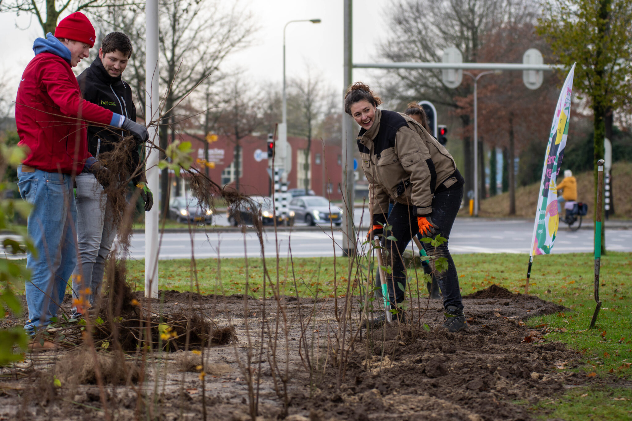 Mensen planten struiken langs een weg met een kleurrijke vlag op de achtergrond.