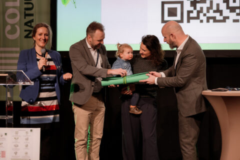Vier mensen en een kind poseren op een podium met een groene tube in een ceremonie.
