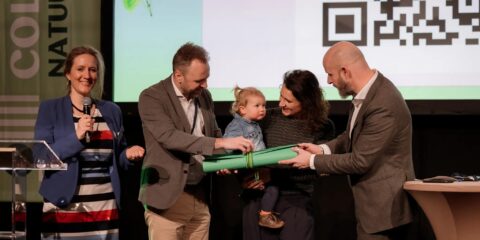 Een vrouw spreekt, terwijl mensen een document uitwisselen op een podium tijdens een evenement.