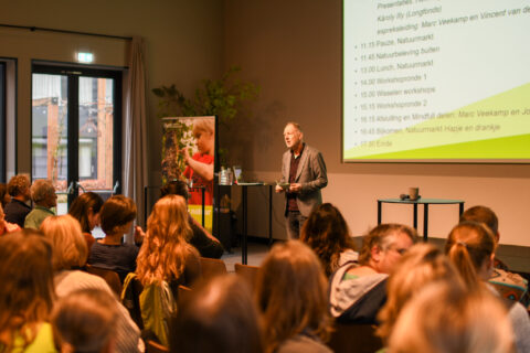 Een spreker presenteert voor een zittend publiek, met een projectiescherm en poster op de achtergrond.
