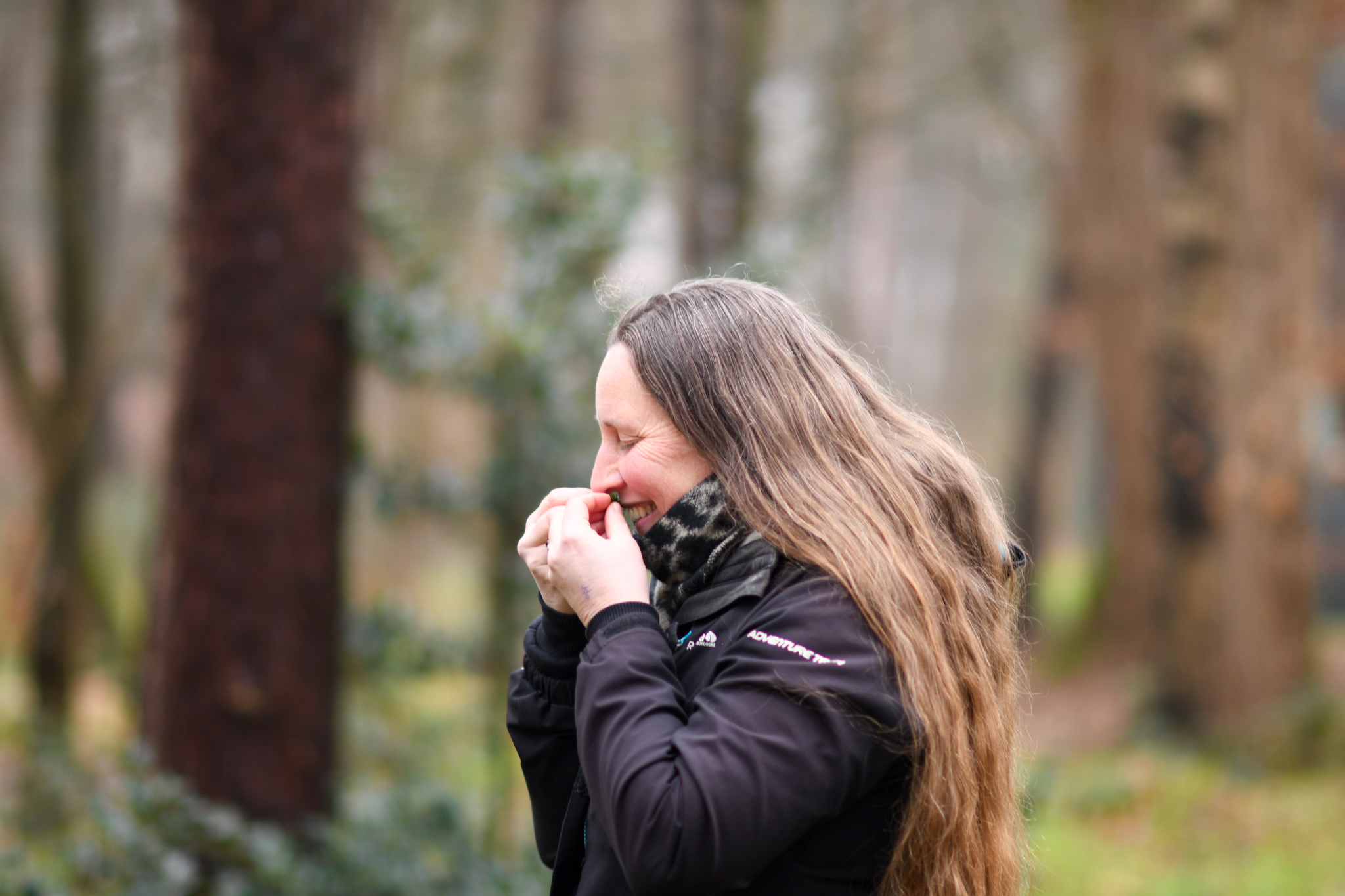 Vrouw met lang haar buiten in een bos, gekleed in een jas, houdt haar gezicht warm met haar handen.