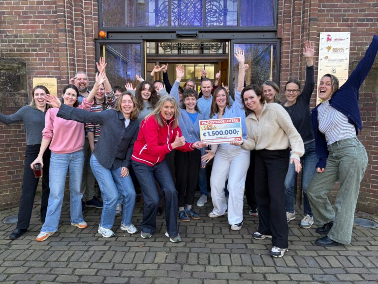 Groep lachende mensen buiten een gebouw, houdt een cheque van €1.500.000 vast.