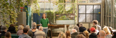 Persoon geeft presentatie over landbouw en natuur in kas gevuld met publiek.