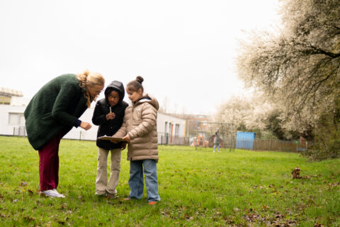 Vrouw en twee kinderen kijken naar een clipboard in een groene tuin met bloesembomen.