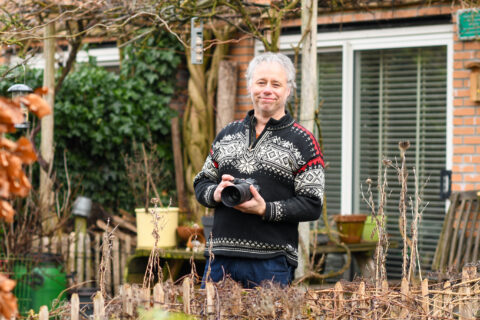 Man in tuin met gebreide trui en camera, omringd door planten en een huis op de achtergrond.