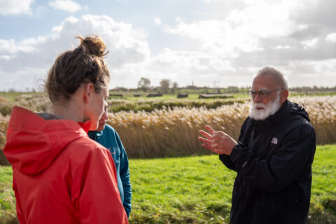 Twee mensen luisteren naar een oudere man die buiten in een landschap iets uitlegt.