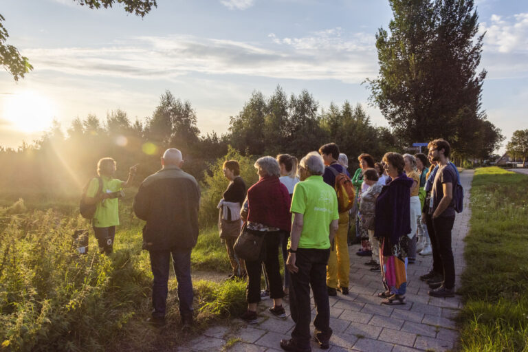 Groep mensen luistert naar gids in groen shirt tijdens zonsondergang op een pad langs een veld.