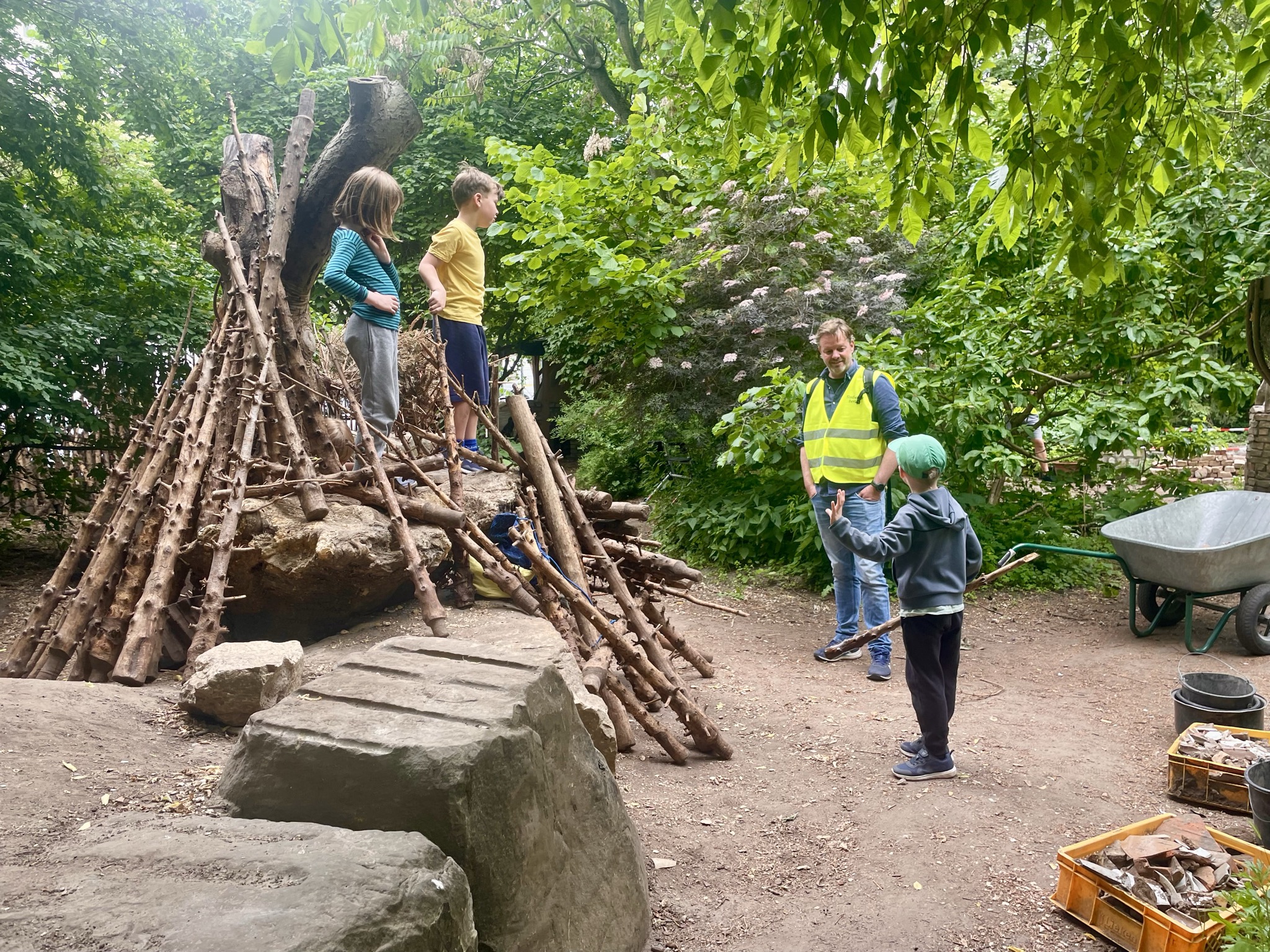 Kinderen spelen bij een houten hut in een bos met een volwassen begeleider in veiligheidshesje.