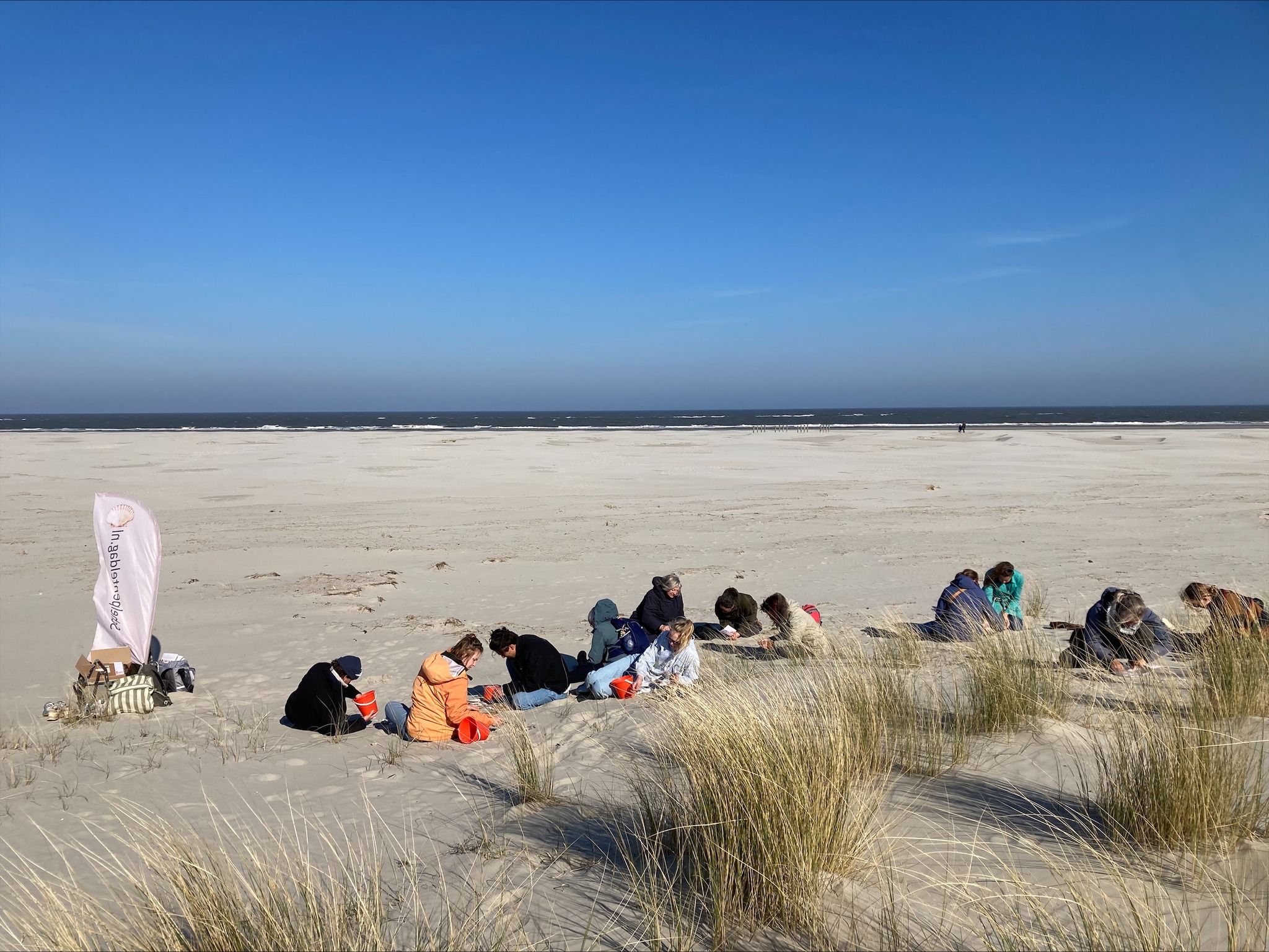 Groep mensen zit op een duin bij de zee met gras en een vlag op de achtergrond.