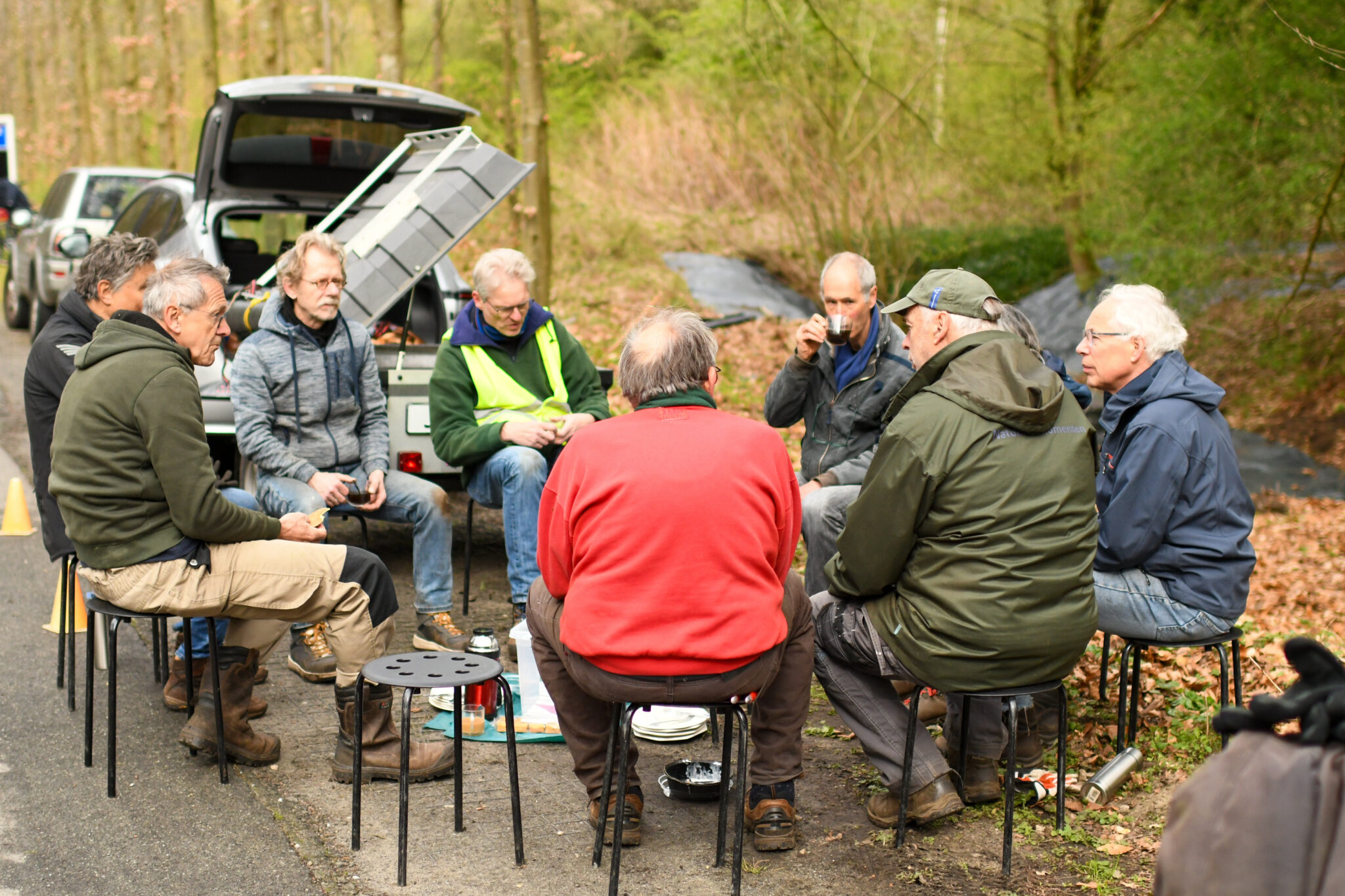 Een groep mannen zit buiten in een kring op krukjes, met een auto en bos op de achtergrond.
