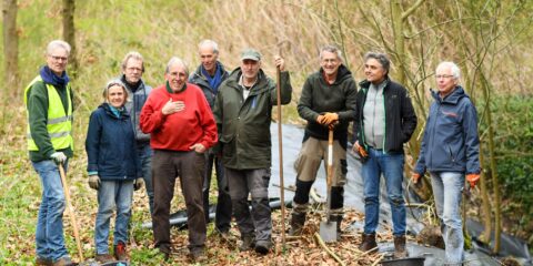 Een groep mensen in werkkleding poseert met tuingereedschap in een bosrijke omgeving.