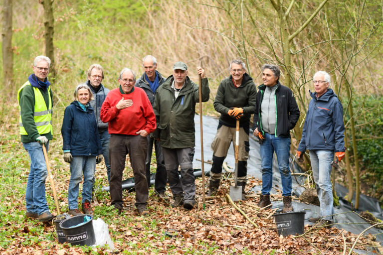 Groep vrijwilligers met tuingereedschap aan het werk in een bosrijke omgeving.