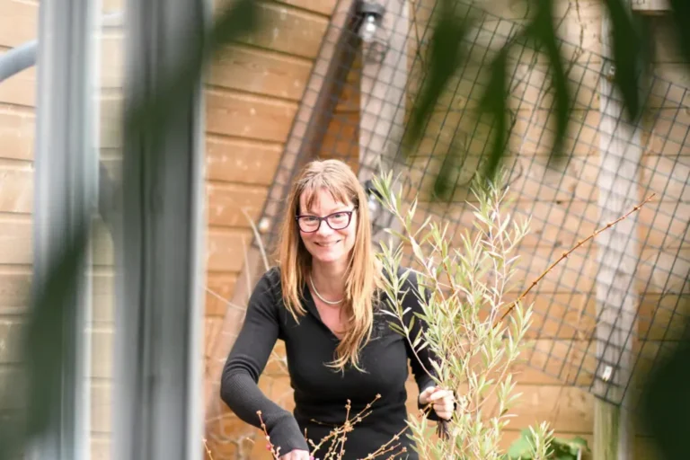Vrouw met bril in tuin, omringd door planten, houten schutting op achtergrond.