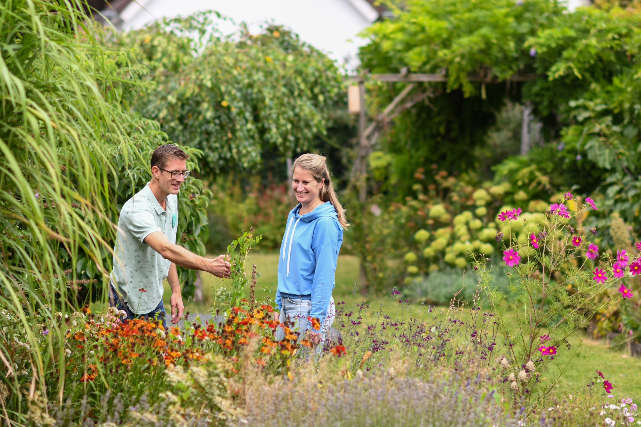Twee personen glimlachen in een kleurrijke tuin met bloemen en groen om hen heen.