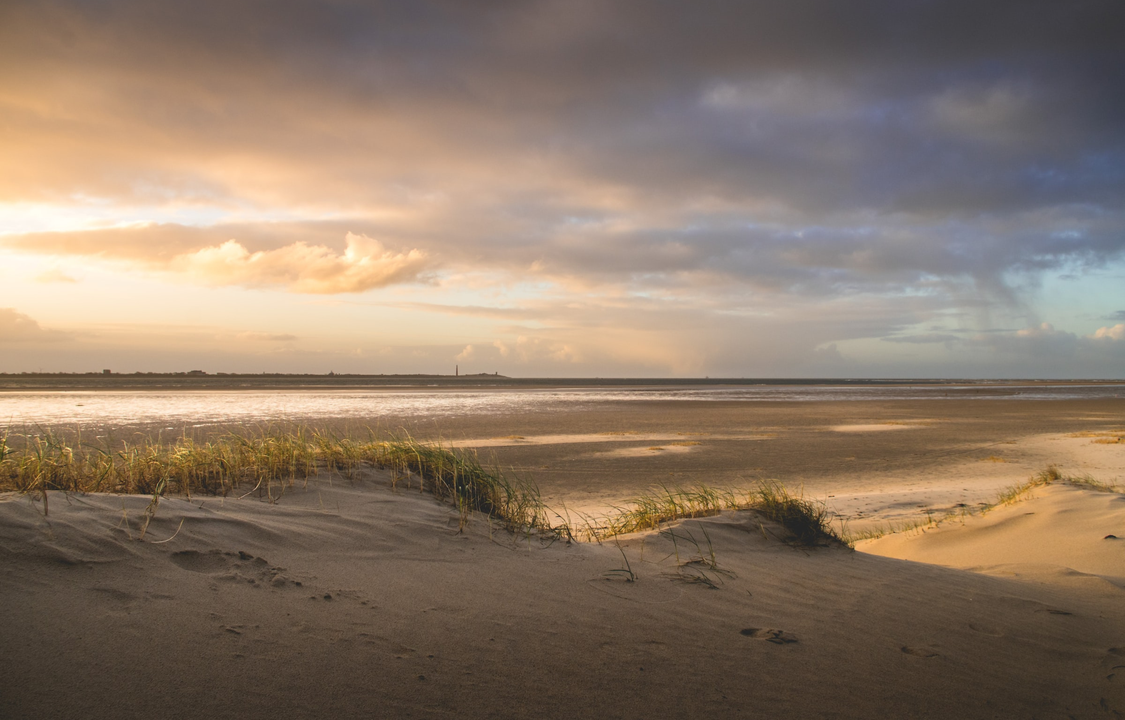 Strandlandschap met grasbedekte duinen bij zonsondergang, donkere wolken en een kalme zee.