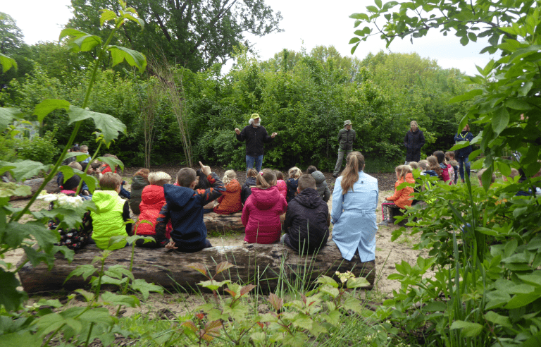 Groep kinderen luistert naar buitenles in bosrijke omgeving.
