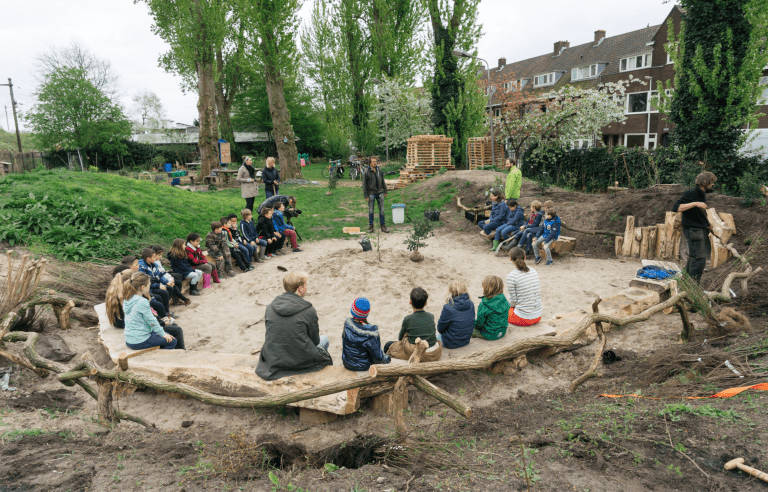 Groep kinderen zit in een kring buiten op boomstammen, omringd door bomen en huizen.