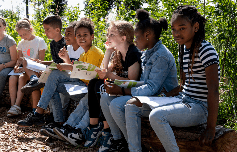 Kinderen zitten buiten op boomstammen en maken aantekeningen in werkboekjes.