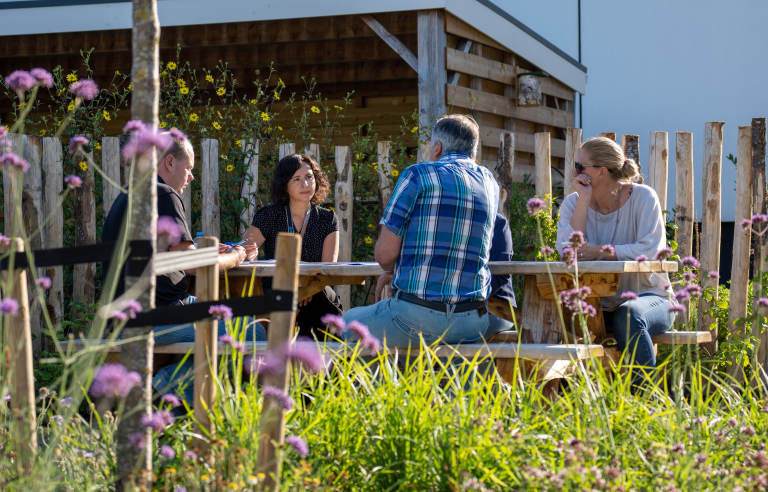 Groep mensen vergadert aan een buitentafel omringd door wilde bloemen en planten.
