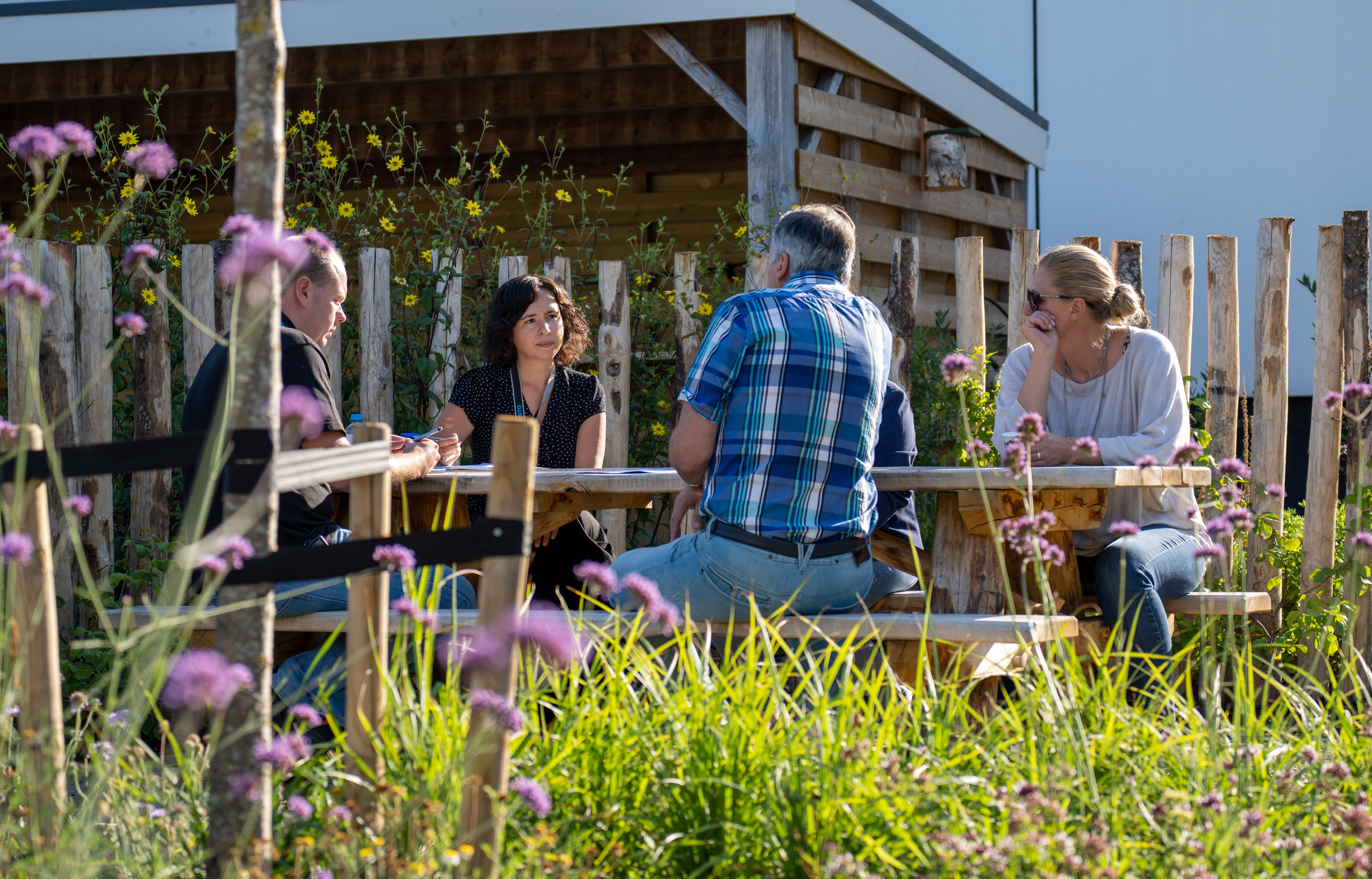 Groep mensen vergadert aan een buitentafel omringd door wilde bloemen en planten.