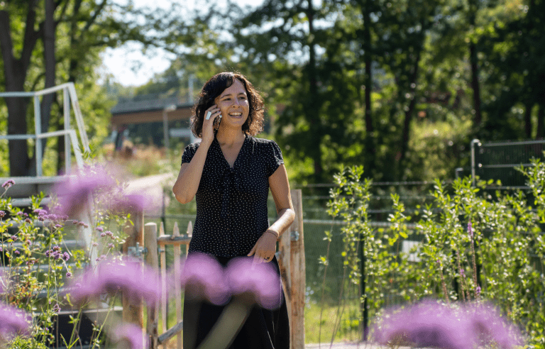 Vrouw telefoneert lachend in een groene, zonnige tuin met paarse bloemen op de voorgrond.